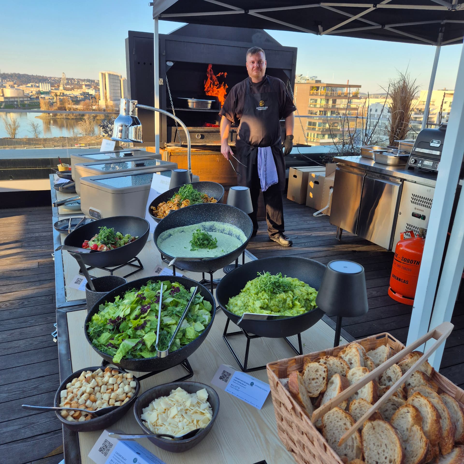 Grill station with salads, sides and bread on a rooftop terrace, a chef working at an open grill behind